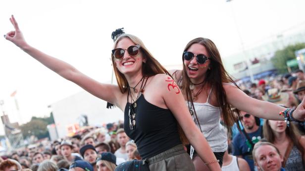 Zwei junge Frauen mit Sonnenbrillen und Körperbemalung feiern auf einem Festival vor einer Menschenmenge.