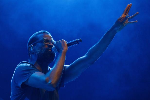 Ein Mann mit Brille singt in ein Mikrofon und hebt eine Hand in der blauen Beleuchtung.