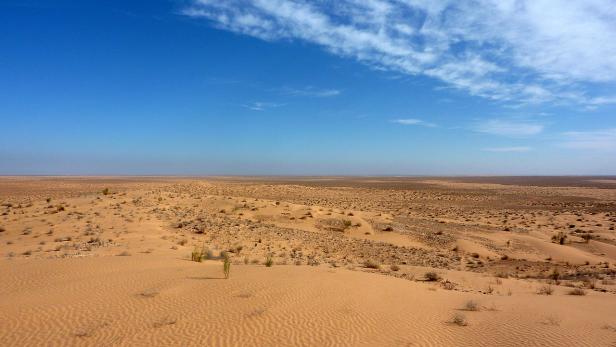 Eine weite, sandige Wüstenlandschaft unter einem blauen Himmel mit vereinzelten Wolken.