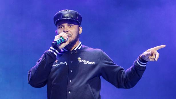 Ein Mann mit Mütze singt in ein Mikrofon und zeigt mit dem Finger nach rechts.