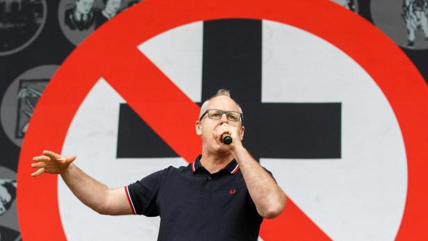 Ein Mann mit Brille singt vor einem durchgestrichenen Kreuz auf einer Bühne.