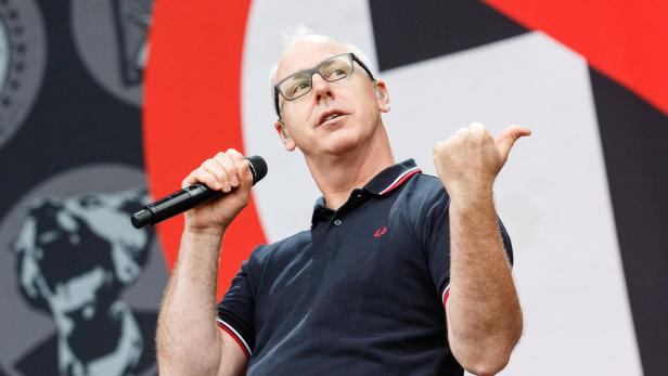 Ein Mann mit Brille und Poloshirt spricht in ein Mikrofon und zeigt mit dem Daumen nach rechts.