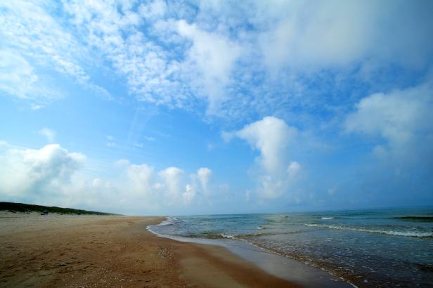 Ein Sandstrand trifft auf das ruhige, blaue Meer unter einem bewölkten Himmel.