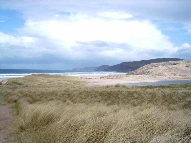 Dünenlandschaft mit Strand und Bergen unter bewölktem Himmel.