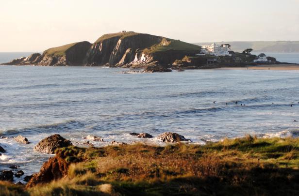 Blick auf eine Küstenlandschaft mit einem Hügel, einem Hotel und Surfern im Wasser.