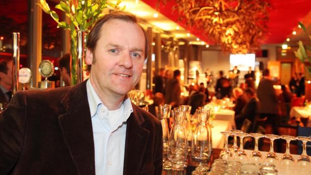 Ein Mann in einem braunen Sakko steht vor einem festlich gedeckten Tisch in einem Restaurant.