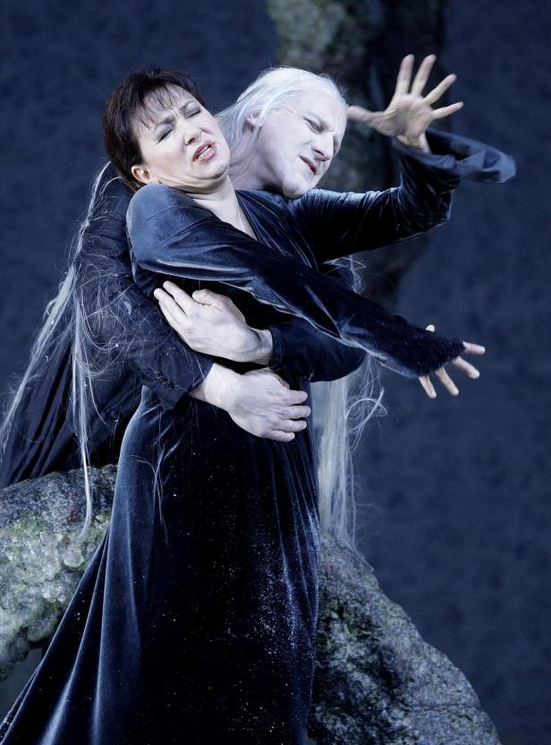 Eine Frau in einem dunklen Kleid wird von einer Figur mit weißer Haut und langen weißen Haaren umarmt.