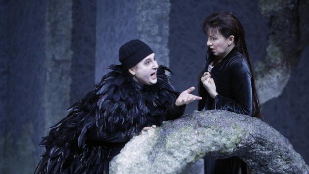 Ein Mann im Krähenkostüm gestikuliert auf der Bühne mit einer Frau im schwarzen Kleid.