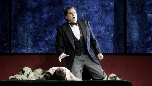 Ein Opernsänger steht über einer liegenden Frau, umgeben von weißen Blumen.