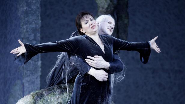 Eine Frau in einem schwarzen Kleid wird von einer weißhaarigen Gestalt umarmt, beide mit schmerzverzerrten Gesichtern.