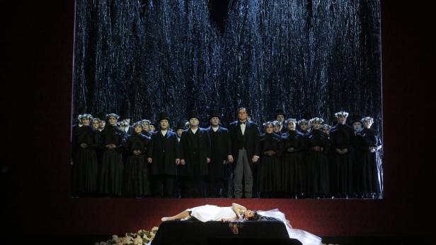 Eine Theaterszene mit einer Frau in Weiß, die aufgebahrt liegt, und einem Chor in Schwarz gekleideter Menschen im Hintergrund.