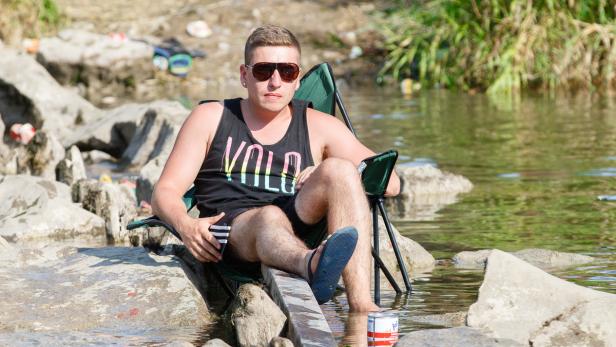 Ein Mann mit Sonnenbrille sitzt auf einem Campingstuhl am Ufer und badet seine Füße im Wasser.