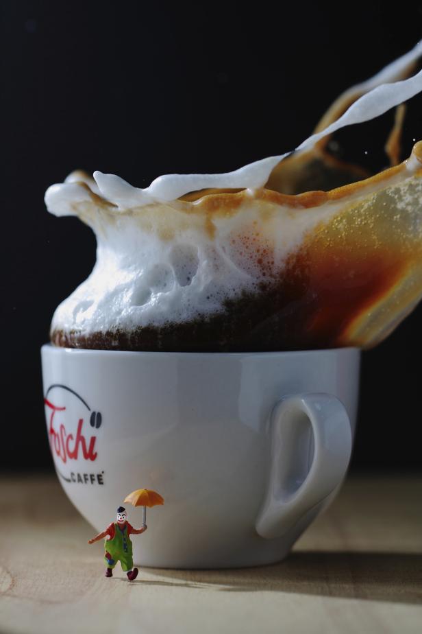 Ein Clown mit Regenschirm steht vor einer Tasse Kaffee, aus der Schaum herausspritzt.