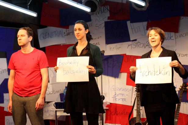 Drei Personen stehen auf einer Bühne und halten Schilder mit Berufsbezeichnungen hoch.