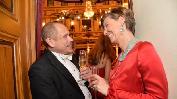 Ein Mann im Smoking und eine Frau im roten Kleid unterhalten sich bei einem Glas Sekt.