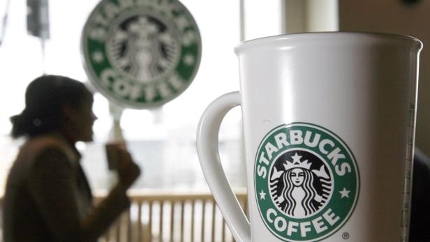 Eine Starbucks-Tasse steht im Vordergrund, im Hintergrund eine Kundin und das Starbucks-Logo.