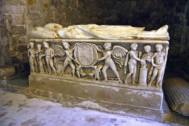 Ein reich verzierter Sarkophag mit einer liegenden Figur und Reliefs von Putten und Wappen.