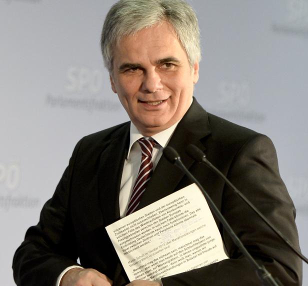 Ein Mann in einem Anzug steht vor Mikrofonen und hält ein Dokument in der Hand.