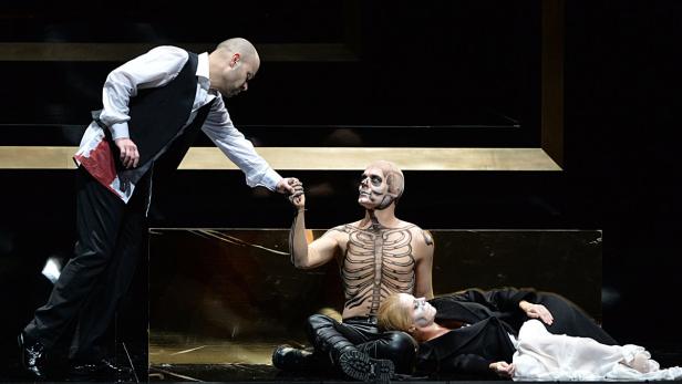 Auf einer Bühne hält ein Mann einen anderen mit Skelett-Bemalung an der Hand, während eine Frau am Boden liegt.