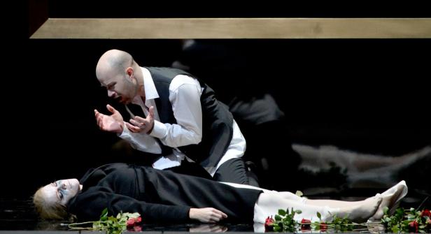 Ein Mann kniet neben einer leblosen Frau, umgeben von roten Rosen.