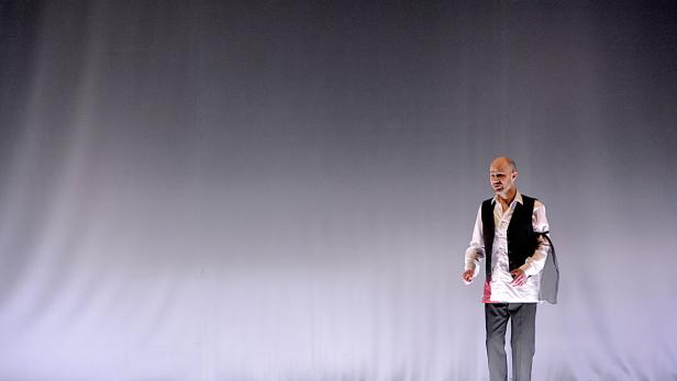 Ein Mann mit Weste steht auf einer Bühne vor einem grauen Hintergrund.