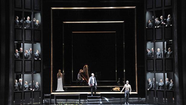 Eine Bühnenszene mit Schauspielern in Kostümen vor einem schwarzen Bühnenbild mit goldenen Rahmen.