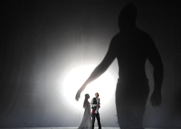 Eine Frau und ein Mann stehen vor einem hellen Licht, überragt von einem großen Schatten.