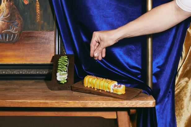 Eine Hand streut Sesam über eine Sushi-Rolle mit Avocado auf einem Holztisch.