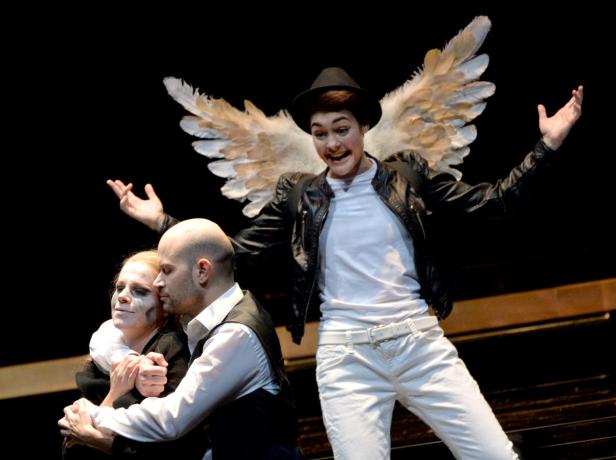 Eine Theaterszene mit einem Mann mit Engelsflügeln und zwei weiteren Schauspielern.