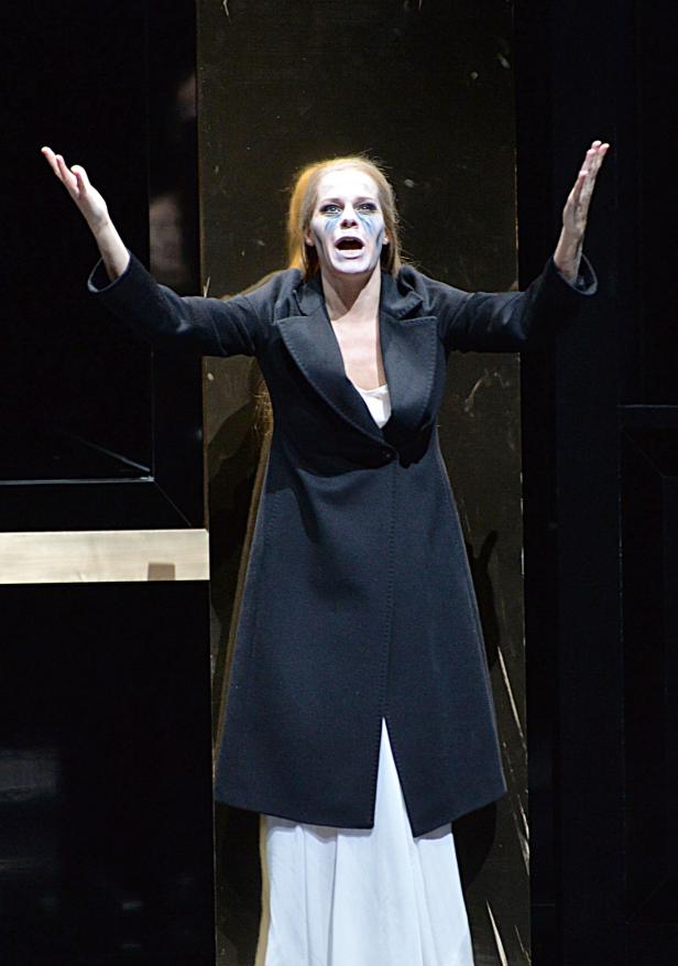 Eine Frau mit theatralischem Make-up und einem schwarzen Mantel steht auf einer Bühne und gestikuliert.