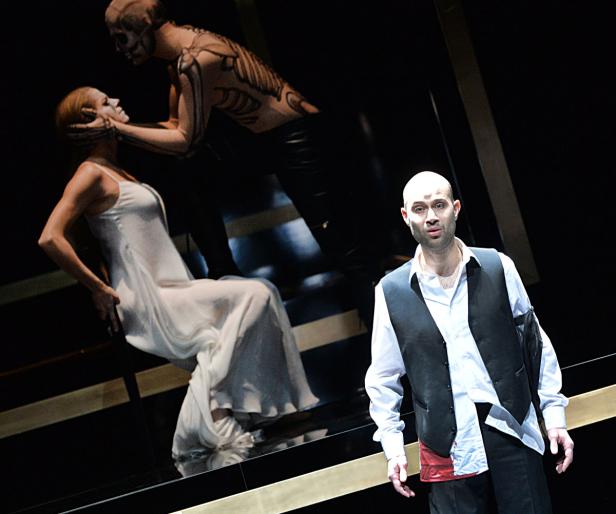 Eine Szene aus einer Opernaufführung mit einem Mann im Vordergrund und zwei Figuren im Hintergrund.