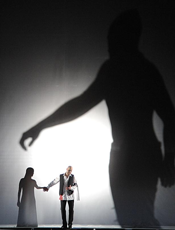 Auf einer Bühne halten ein Mann und eine Frau Händchen, während riesige Schatten auf eine Leinwand projiziert werden.
