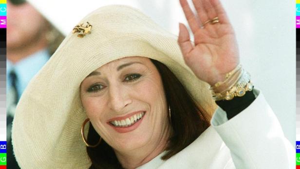 Anjelica Huston mit Hut winkt lächelnd in die Kamera.