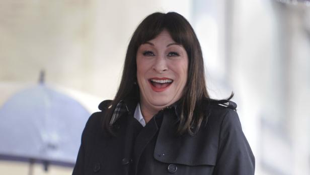 Anjelica Huston lächelt in einem schwarzen Mantel.