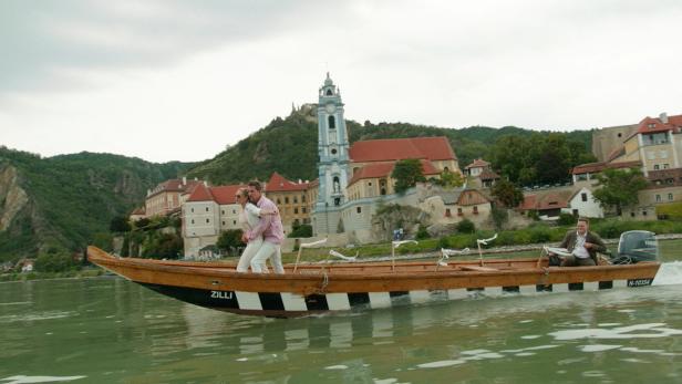 Ein Paar steht in einem Boot auf der Donau, im Hintergrund die Stadt Dürnstein mit der Stiftskirche.