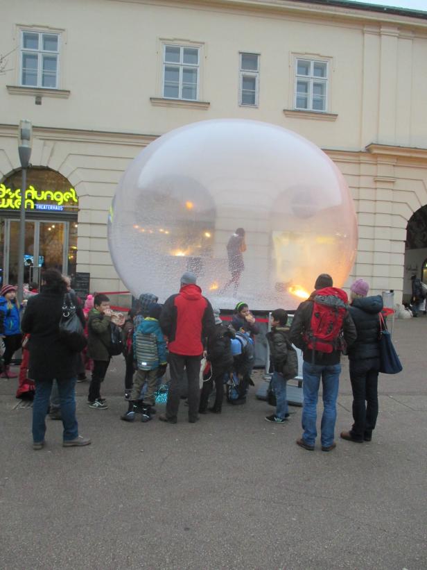 Eine Gruppe von Menschen steht vor einer großen, transparenten Schneekugel vor dem Theaterhaus Dschungel Wien.