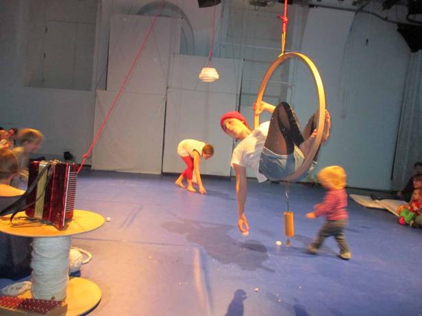 Eine Frau hängt in einem Reifen von der Decke, während Kinder auf dem Boden spielen.