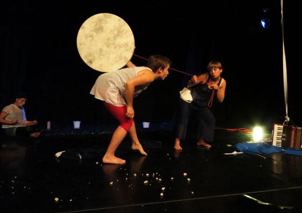 Zwei Performer interagieren auf einer Bühne mit einem großen, runden Objekt und einer roten Schnur.