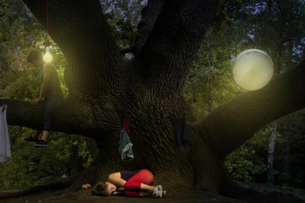 Eine Frau schläft unter einem großen Baum, während eine andere auf einem Ast sitzt; Kleidung hängt am Baum.