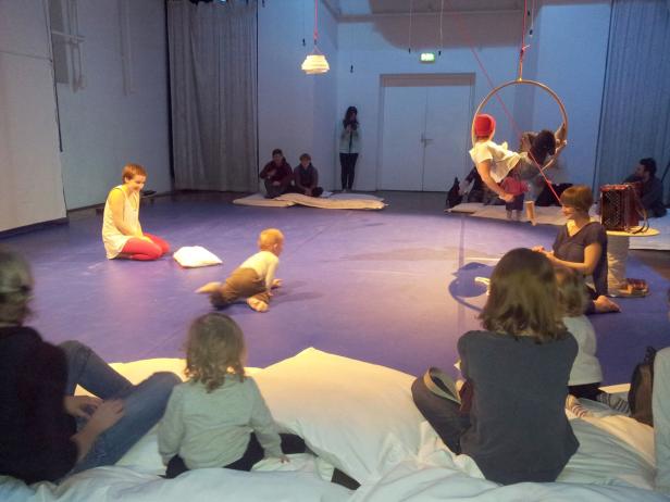 In einem Raum sitzen und liegen Menschen auf Matten und Kissen, während ein Kind krabbelt und eine Person in einem Reifen hängt.