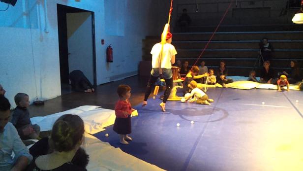 Eine Artistin hängt an einem Seil, während Kinder auf dem Boden sitzen und zusehen.
