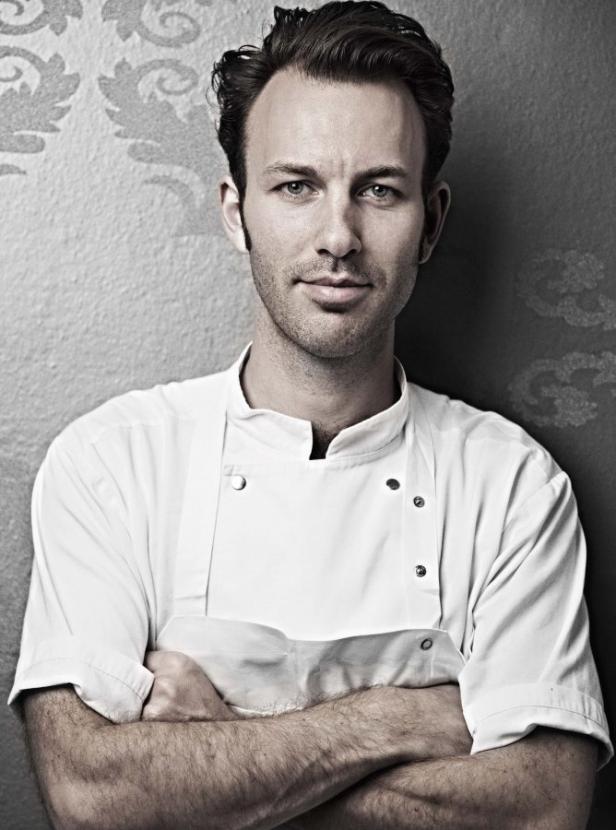 Ein Mann in Kochkleidung steht mit verschränkten Armen vor einer Wand.