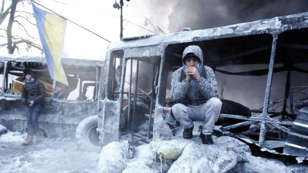 Ein Mann kauert vor einem ausgebrannten Bus mit ukrainischer Flagge.