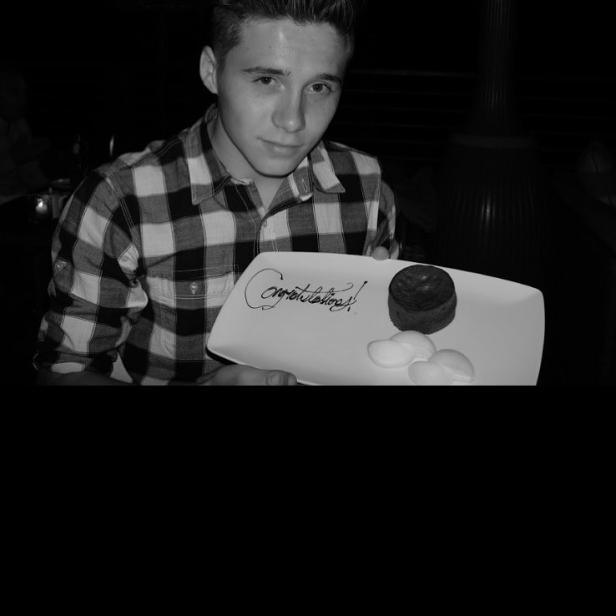 Ein junger Mann hält einen Teller mit einem Schokoladenkuchen und der Aufschrift „Congratulations!“.