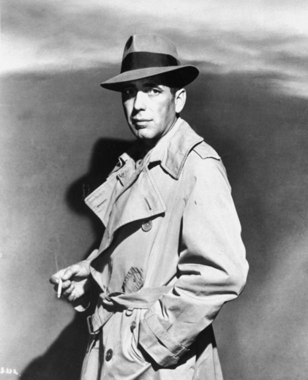 Humphrey Bogart mit Trenchcoat und Hut, eine Zigarette in der Hand.