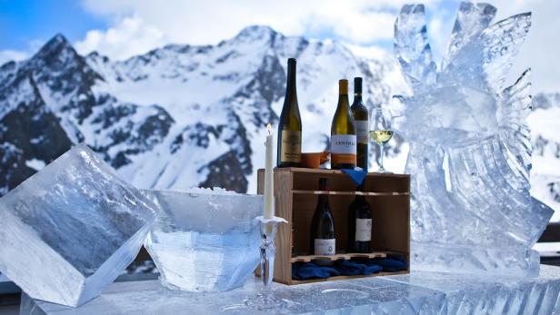Eisskulpturen und Weinflaschen vor einer verschneiten Bergkulisse.
