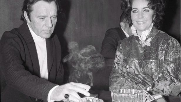 Ein Schwarzweißfoto von Richard Burton, der raucht, und Elizabeth Taylor, die lächelt.