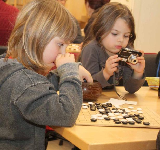 Zwei Kinder spielen das Brettspiel Go, eines fotografiert mit einer Kamera.