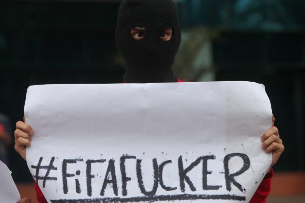 Eine Person mit Sturmhaube hält ein Schild mit der Aufschrift „#FIFafucker“.