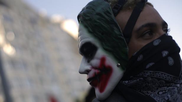 Eine Person mit einer „Joker“-Maske und einem schwarzen Bandana vor einem Gebäude.